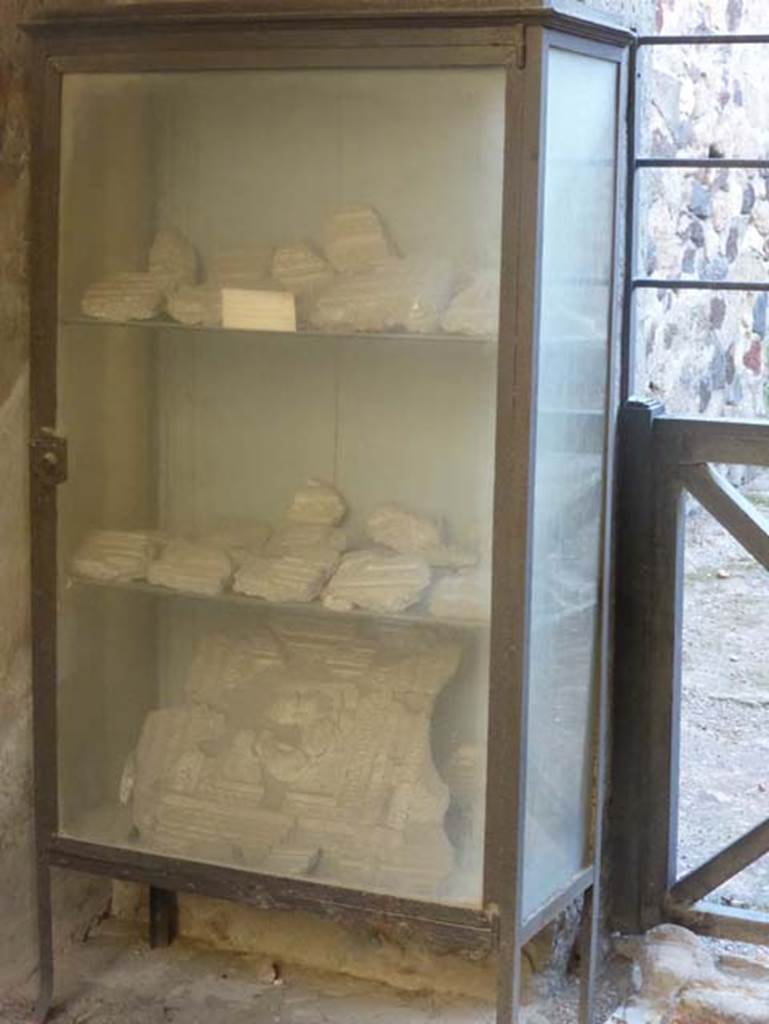 VI.16 Herculaneum. September 2015. One of the ancient glass display cabinets placed at Herculaneum by Maiuri.  Now containing some fragments of stucco belonging to the four-sided arch opposite the shop, on the north side of Decumanus Maximus.
