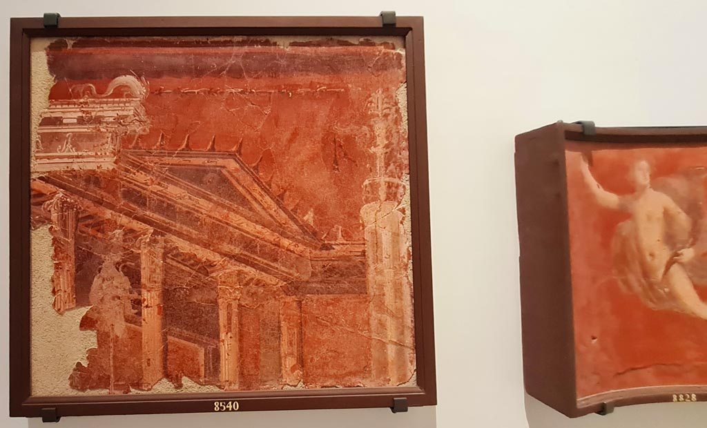 Herculaneum Augusteum. April 2023. Paintings on wall to left of Arch through to display of items from the Theatre. 
On left – Building with tympanum on a red background – inv. 8540. Photo courtesy of Giuseppe Ciaramella.
