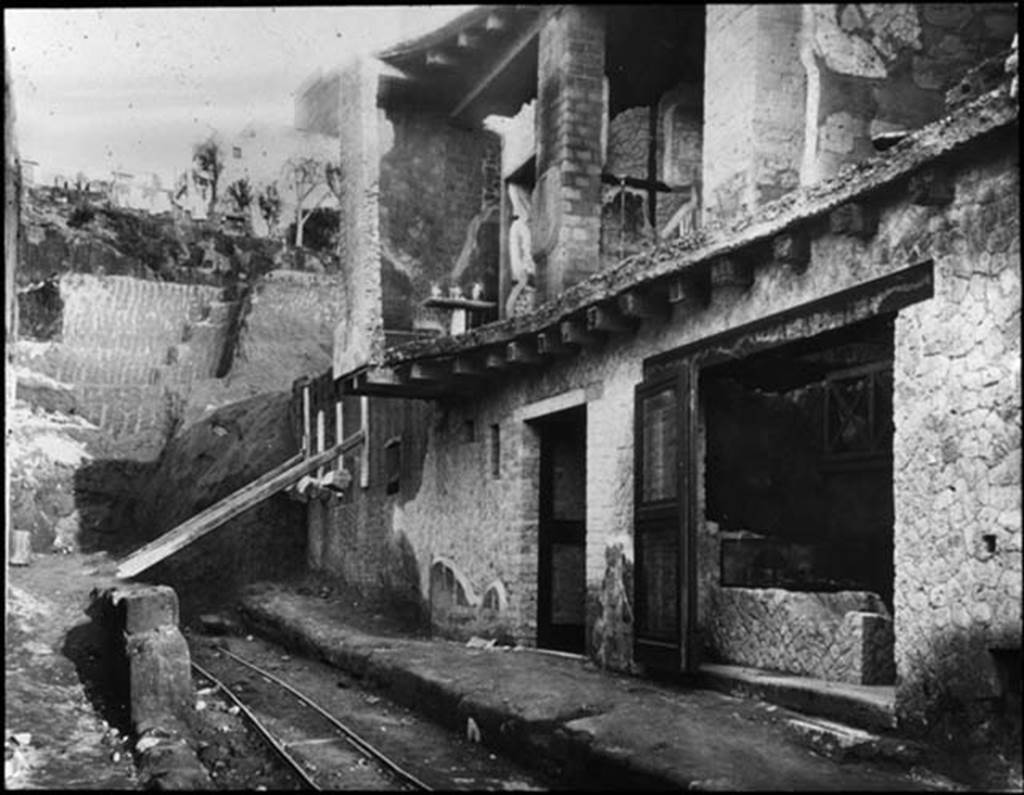 Cardo IV Superiore, Herculaneum. Looking north along Cardo IV Superiore towards Insula V., during excavations.1935.  Photo by Fratelli Alinari (I.D.E.A.). Alinari No 43312.  V.6, the doorway to the wine/food shop is on the right.  V.7, centre doorway with an upper floor is Casa di Nettuno e Anfitrite or House of Neptune and Amphitrite.  V.8, is the area still being excavated beneath props holding wall up, on left.  Used with the permission of the Institute of Archaeology, University of Oxford. File name instarchbx116im012 Resource ID 42241.
