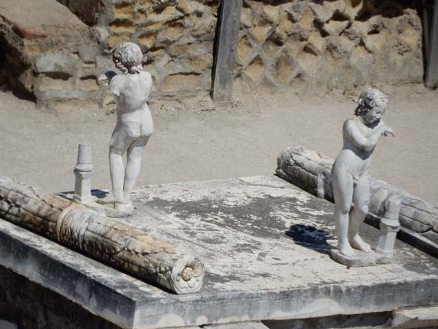 Herculaneum, May 2018. Two marble statuettes on top of altar. Photo courtesy of Buzz Ferebee.
According to Jashemski – “The beautiful statuette of the boy Eros found on the terrace below (that is below and belonging to The House of the Stags) together with the fragments of another statuette of the same subject are believed to have belonged to the same collection of art”.
See Jashemski, W. F., 1993. The Gardens of Pompeii, Volume II: Appendices. New York: Caratzas. (p.265).
According to Deiss, “another statuette found on the terrace [of the House of the Stags] of extraordinary quality is the “Boy Eros”, a nude adolescent with swirling curls (once painted red) and dreamy face.”
See Deiss J. J., 1968. Herculaneum: A city returns to the sun. London: History Book Club, p. 43. 
