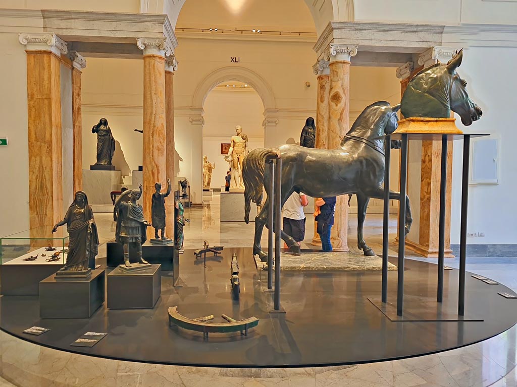 Herculaneum, April 2023. Bronzes in “Campania Romana” gallery in Naples Archaeological Museum. 
Looking across gallery towards arch to gallery with items from Theatre, and through another Arch towards items from Augusteum.
On the left in the glass case are the small bronze chariot attachments. Photo courtesy of Giuseppe Ciaramella.
