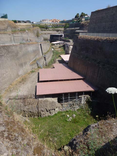 Villa dei Papiri complex, June 2014. Overview of site, looking west.
Photo courtesy of Michael Binns.
From front to back are:
The double roof of the House of the Dionysiac Reliefs, seaside pavilion.     
The paler coloured roof around the corner is the House of the Dionysiac Reliefs.
The tall building of the baths.
The white roof covering the collapsed monumental structure.
The levels of the Villa dei Papiri itself.

